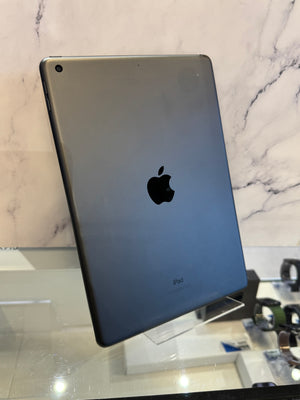 An Apple iPad with a dark gray metallic back, standing on a clear display stand on a glass table against a marble-patterned wall.