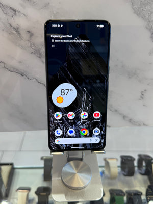 A black Pixel smartphone displayed upright on a metallic stand in a retail setting, with a marble-patterned background and watch displays blurred below.