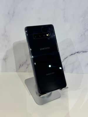 A black Samsung Galaxy S10+ smartphone shown from the back, resting on a clear display stand against a white marble-patterned background.