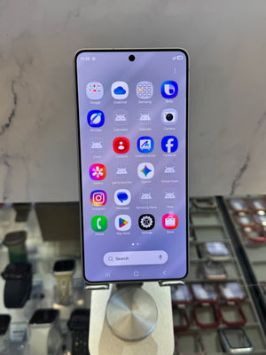 A Samsung Galaxy smartphone with a black front bezel and a colorful app grid on the screen, displayed on a silver stand in a retail setting.