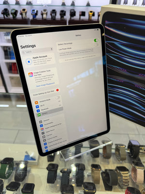 An iPad displaying the Settings app on a clear acrylic stand, with a watch display case and other smartwatches visible in the blurred background of a retail store.