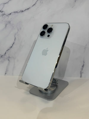 A silver Apple iPhone 12 Pro shown from the back, resting on a gray metal phone stand against a white marble-patterned background.