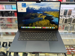 A dark gray Apple MacBook laptop with an open screen displaying a mountain landscape wallpaper, set on a glass display counter in a retail store.