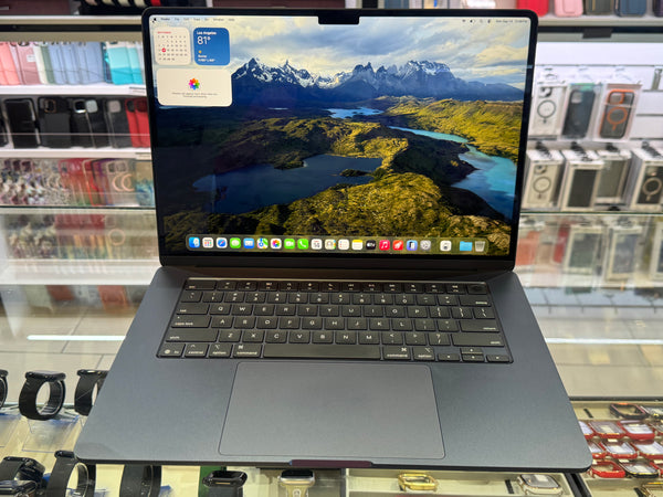 A dark gray Apple MacBook laptop with an open screen displaying a mountain landscape wallpaper, set on a glass display counter in a retail store.