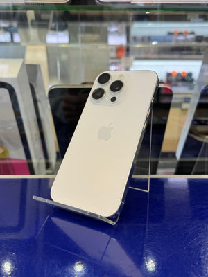 A white Apple iPhone shown from the back, resting on a clear acrylic stand inside a retail display case with a blue wall in the background.