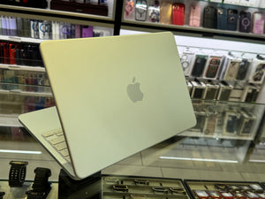 A silver Apple MacBook laptop shown open at an angle, resting on a glass display counter inside a retail store. The Apple logo is clearly visible on the back of the laptop lid, with shelves of phone cases and accessories blurred in the background.
