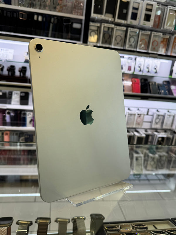 A silver Apple iPad shown from the back, resting on a clear display stand inside a retail store with shelves of other electronics in the blurred background.