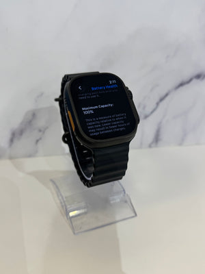 A black smartwatch with a rectangular screen and black band, displayed on a clear stand against a white marble-patterned background.