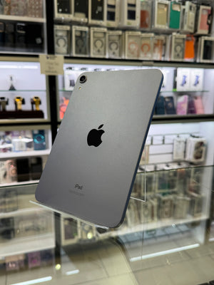 A silver Apple iPad shown from the back, resting on a clear display stand inside a retail store with shelves of phone accessories in the background.