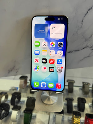 A black iPhone with its screen on, displaying the home screen, resting on a silver metal stand. The background shows a marble-patterned wall and blurred watch displays on a counter.