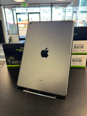 A silver Apple iPad shown from the back, standing upright on a clear display stand on a dark tabletop. Apple logo and the word 'iPad' are visible on the back, with product boxes in the background.