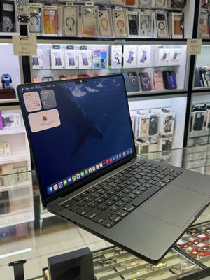 A dark gray Apple MacBook laptop with its screen turned on, displayed on a glass counter inside a retail store with shelves of phone accessories in the background.