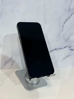 A black smartphone with a blank screen stands upright on a silver metal adjustable phone stand against a white marble-patterned background.