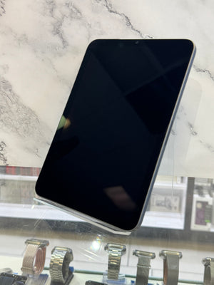 A black tablet with rounded corners displayed at an angle on a stand, set against a white marble-patterned background with watch displays visible below.