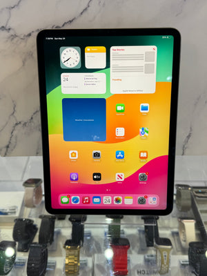 An iPad with a colorful gradient home screen, standing upright on a glass display case with various smartwatches in the background.