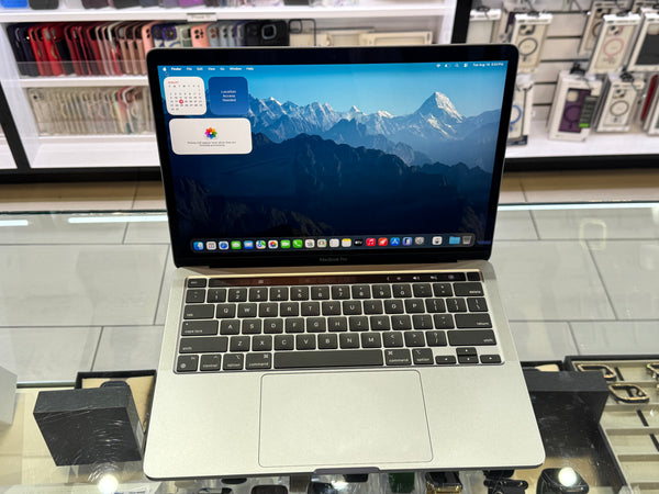 A silver Apple MacBook laptop with a black screen border and keyboard, displayed open on a glass counter in a retail store setting.