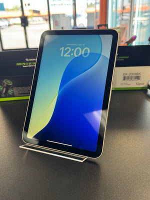 A black tablet with a clear acrylic stand on a dark tabletop, showing a blue and green lock screen wallpaper. The background shows a blurred store window and packaging boxes.