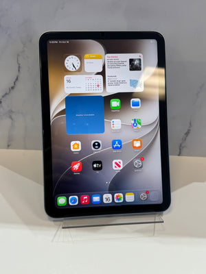 A black iPad Pro tablet on a clear acrylic stand, displaying its home screen with various app icons against a light gray marble-patterned background.