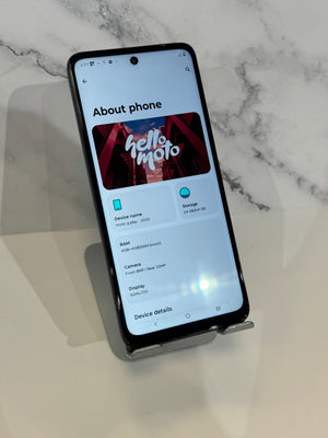 A black smartphone displayed upright on a small stand, showing the 'About phone' screen with Samsung branding. The phone is photographed on a light marble-patterned surface with a soft shadow.