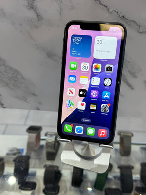 A black smartphone with a lit home screen displayed on a white stand against a marble-patterned background.