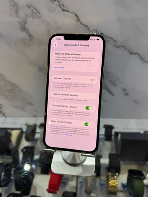 A black smartphone on a metal display stand showing a pink battery health charging screen, set against a marble-patterned wall and blurred watch display.