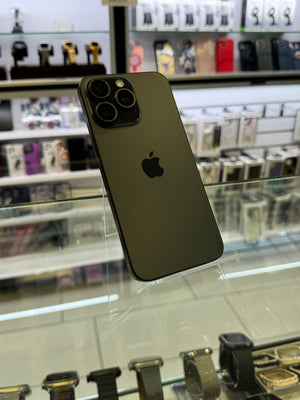 A dark green Apple iPhone Pro showing the back of the device with its triple-camera system and the Apple logo, displayed on a glass stand inside a retail store.