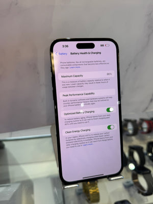 A black smartphone displaying the iPhone Battery Health & Charging settings screen, standing on a small display stand with a blurred watch and countertop in the background.
