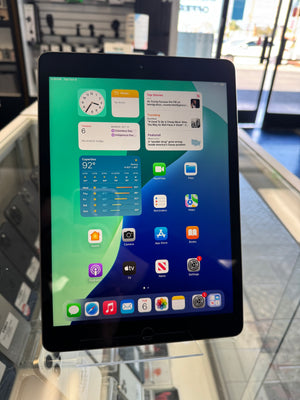 A black Apple iPad tablet displayed on a glass stand inside a retail store, showing the home screen with various app icons.