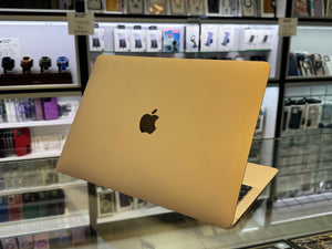 A gold Apple MacBook laptop is shown open on a glass display counter inside a retail store, with shelves of accessories and electronics blurred in the background.