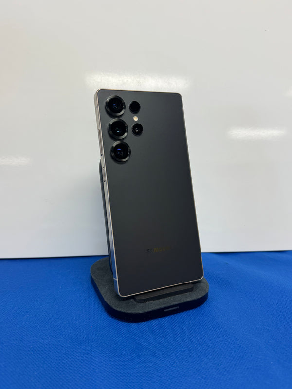 A dark gray Samsung Galaxy Ultra smartphone shown from the back, resting on a black charging stand against a white background with a blue surface below.