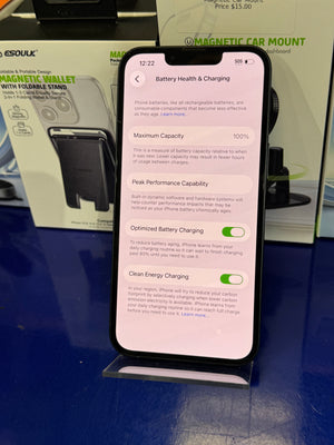 Smartphone displaying battery health and charging settings on a reflective surface with product packaging in the background.