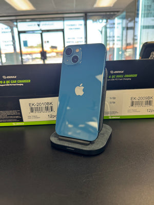 A blue Apple iPhone stands upright on a charging dock on a dark tabletop, with two black and green Esuke charger boxes behind it in a retail or office setting.