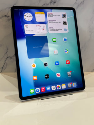 A tablet device with a black bezel and silver edges, shown standing upright on a clear stand against a light marble-patterned background. The screen is on and displaying colorful app icons and widgets.