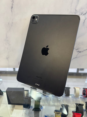 A black Apple iPad Pro shown from the back, standing on a clear display stand in a retail or showroom setting with marble wall panels and other devices visible below.