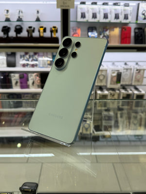 A Samsung smartphone shown from the back, resting on a glass display case in a retail store. The phone has a pale mint green back panel with a dark blue-gray frame and a large multi-camera system on the top left.