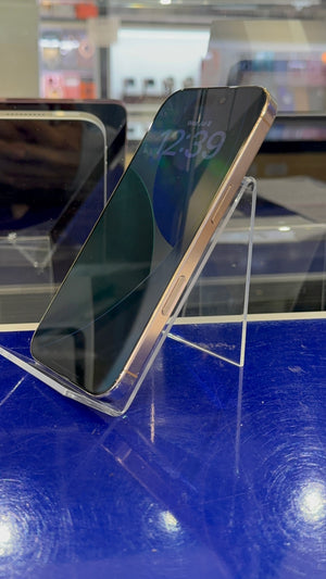 A smartphone with a dark screen and a metallic rose gold or light gold frame, displayed on a clear acrylic stand on a blue reflective surface.