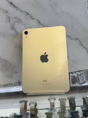 A gold-colored iPad showing its back panel, featuring the black Apple logo and 'iPad' text, displayed on a stand against a white marble-patterned background.
