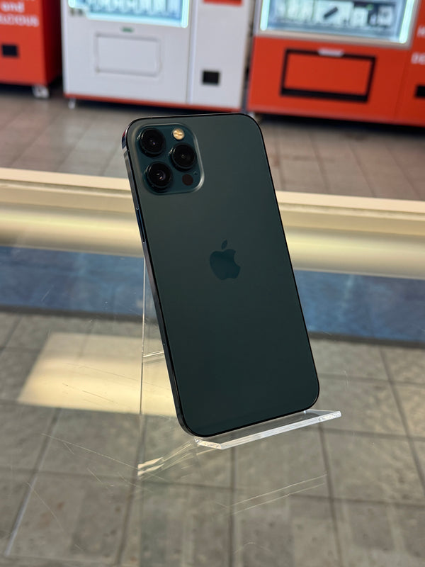 A dark green Apple iPhone 12 Pro shown from the back, resting on a clear acrylic display stand in a store setting.
