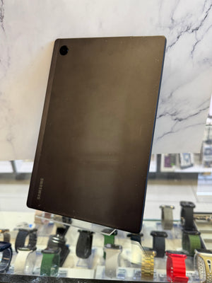 A Samsung-branded black rectangular accessory, likely a protective skin or cover, displayed on a white marble-patterned surface with a blurred watch display case in the background.