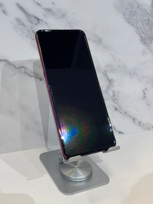 A black smartphone with a red side frame stands upright on a silver metal stand against a white marble-patterned background.