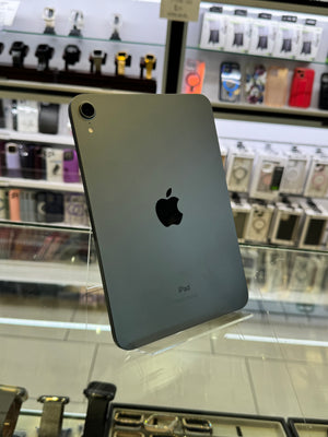 A grey Apple iPad shown from the back, resting on a glass display counter inside a retail store with shelves of accessories blurred in the background.