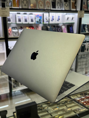 A silver Apple MacBook laptop shown open at an angle, with the Apple logo visible on the back of the lid. The laptop is photographed inside a retail store with display shelves and products blurred in the background.