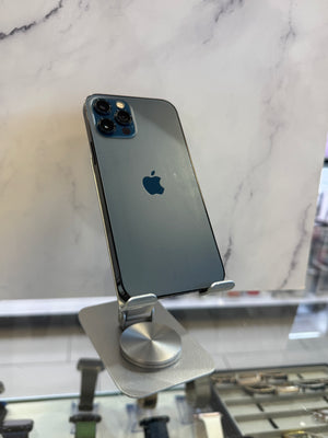 A dark blue-gray Apple iPhone 12 Pro shown from the back, mounted on a silver display stand on a white marble surface.