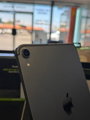 A dark gray Apple iPad shown from the back, featuring a single rear camera lens and the Apple logo, photographed in a store setting with a blurred background.