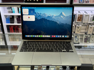 A silver Apple MacBook Pro laptop is open on a display stand inside a retail store, showing a mountain landscape wallpaper and various docked apps on the screen.