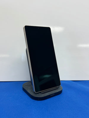 A black smartphone with a silver edge stands vertically on a dark gray charging dock against a white wall, resting on a blue textured surface.