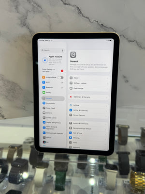 A black tablet device displaying the Settings app on a white marble surface, with blurred watch displays visible in the background.