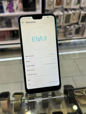 A black smartphone displaying the EMUI about phone screen, resting on a clear acrylic stand on a glass display counter with blurred electronics in the background.