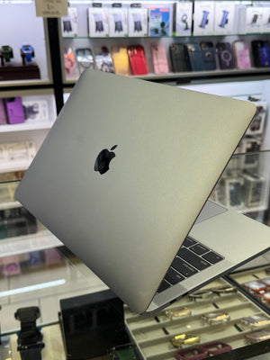 A silver Apple MacBook laptop shown open at an angle, with the Apple logo visible on the back of the lid. The laptop is photographed inside a retail store with display cases and accessories in the background.