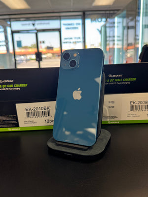 A blue Apple iPhone with a dark gray case stands vertically on a small display stand in front of its retail packaging box. The Apple logo is visible on the back of the phone.